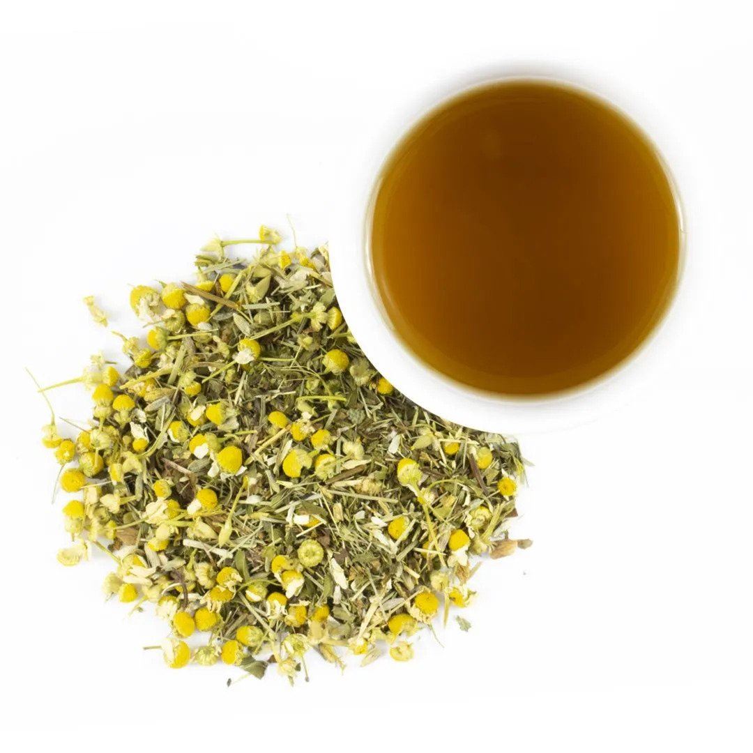 A cup of Daily Defense Herbal Tea rests beside a pile of loose, dried chamomile flowers with yellow centers and pale petals, all set against a white background.